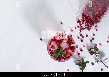 Cold cocktail with lingonberry, ice cubes, straws, scattered berry in transparent glass on soft light white wood table with copy Stock Photo