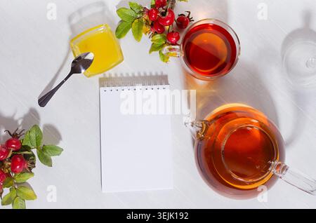 Autumn teatime with rosehip tea with teapot, cup, autumn rose hip branch and blank notepad in sunny day with shadows on white wo Stock Photo