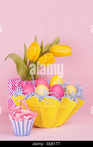 Easter festive background - yellow, blue, red eggs in yellow basket, tulips, cupcake on pastel pink background. Stock Photo