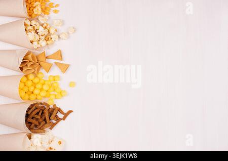 Golden colorful snacks - nachos, popcorn, croutons, chips in craft paper cone as border on white wood table with copy space. Stock Photo