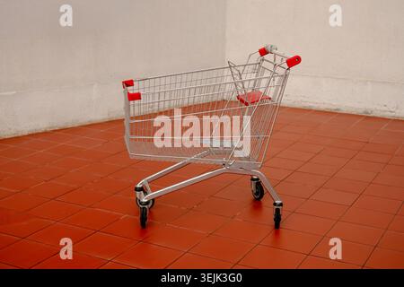 An empty steel supermarket trolley standing alone on an orange tiled floor against a white wall Stock Photo