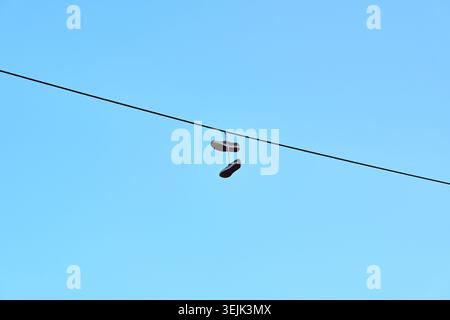 A pair of shoes tied together and hanging from an overhead wire against a blue sky. Stock Photo