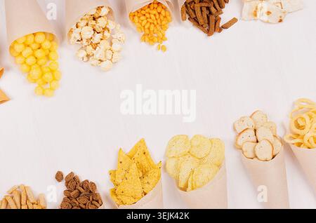 Golden colorful snacks - nachos, popcorn, croutons, chips in craft paper cone as border on white wood table with copy space. Stock Photo