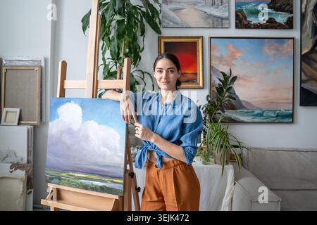 Happy talented female painter posing at camera with finished artwork beside easel at home workshop. Stock Photo