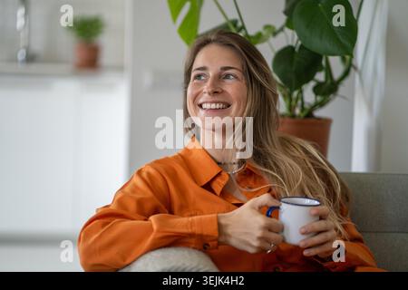 Middle-aged woman with warm smile on face, looking aside while comfortably seated on sofa with cup Stock Photo