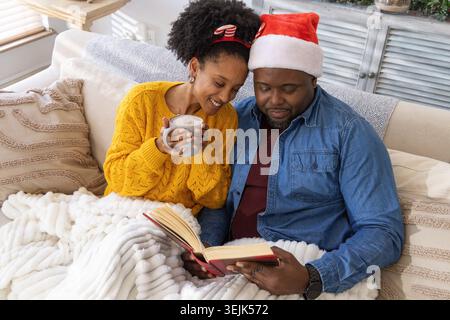 African American couple sitting on sofa at home, man wearing Santa hat, woman holding gray mug. Cozy, holiday, cup, reading, blanket, pillows, knit Stock Photo