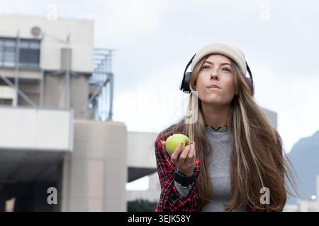 Woman holding green apple, wearing headphones, beanie and red plaid, gazing upward near building. Cityscape, urban, contemplativ Stock Photo