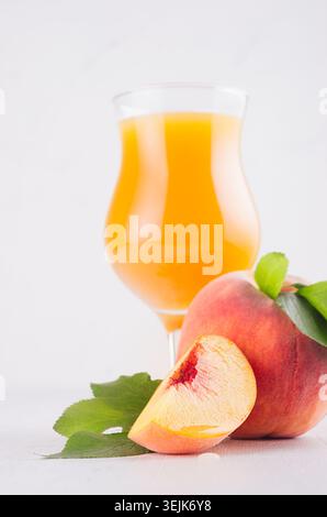 Fresh peach juice in glass with whole peach, ripe juicy slice and green leaves in white modern kitchen interior, closeup, vertic Stock Photo