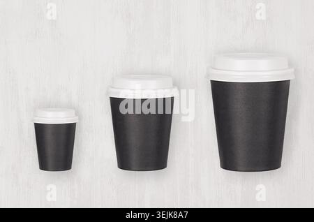 Coffee mockup - set of three different size black paper cups and blank white caps on white wood board, top view. Modern elegant Stock Photo
