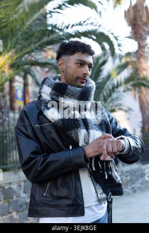 Asian man standing on path among palms wearing leather jacket adjusting watch and holding scarf. Plaid, neckwear, urban, walkway, streetstyle, dayligh Stock Photo