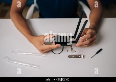 Close-up top view of technician hands assembling wi-fi module on white table, using small screwdriver and other components for computer upgrade or Stock Photo