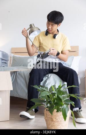 Asian man in his twenties wearing yellow T-shirt inspecting metal desk lamp on bed with headphones Stock Photo
