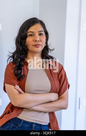 Woman standing with arms crossed, wearing rust shirt, beige tank top and jeans near doorframe Stock Photo