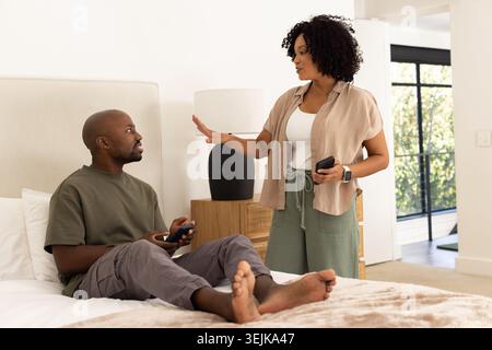 African American couple with woman standing by bed holding phone, man sitting on bed holding remote Stock Photo