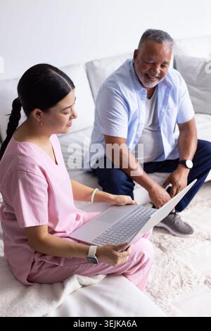 Caregiver in pink scrubs holding silver laptop and guiding senior man on light-colored sofa at home Stock Photo