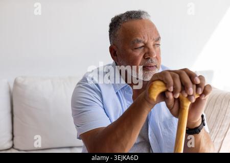 Senior African American man sitting on pale sofa wearing casual shirt and wristwatch holding cane Stock Photo
