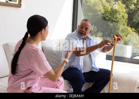 African American senior and Asian caregiver in pink scrubs sitting on couch at home, holding cane Stock Photo