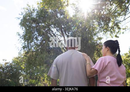 African American man with Asian nurse placing hand on shoulder beside park tree in pink scrubs Stock Photo