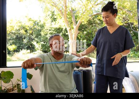 Diverse therapist in scrubs guiding patient doing chest press with blue band by window, copy space Stock Photo