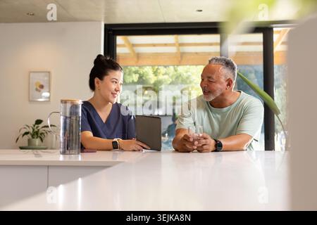 Diverse senior man holding glass and Asian nurse in navy scrubs using tablet at kitchen island Stock Photo