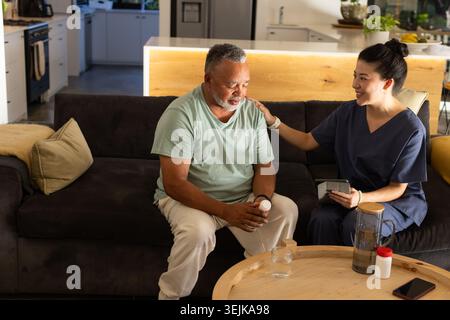 African American senior holding pill bottle while Asian caregiver navy scrubs using tablet on sofa Stock Photo