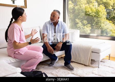 Chinese woman nurse and mature African man sitting on sofa, gesturing and listening with bag nearby Stock Photo