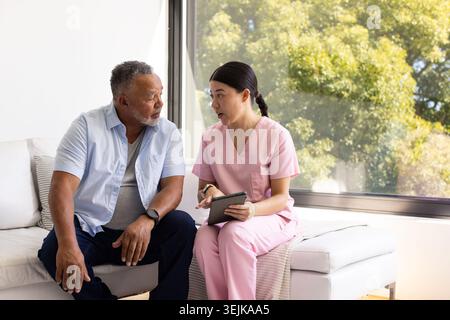 African American man, Asian woman in pink scrubs discussing care using tablet on living room sofa Stock Photo