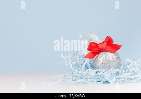 Festive christmas background - silver shiny ball with red ribbon on white wood table and pastel blue wall. Stock Photo