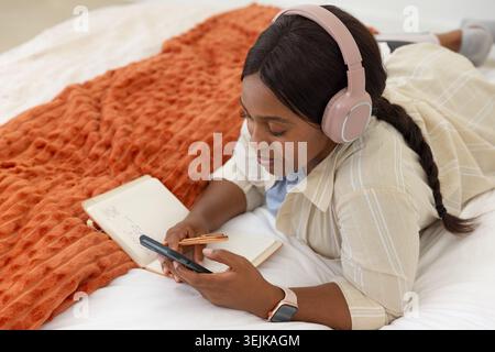 African woman lying on bed writing in notebook while holding pen and using smartphone Stock Photo