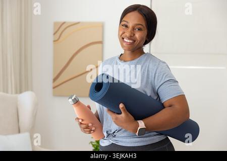 African woman standing in living room holding yoga mat and peach bottle, wearing knotted tee Stock Photo