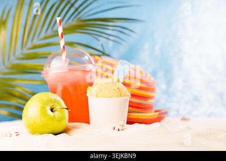 Enjoyment summertime - picnic on tropical beach with cold fruit cocktail, ice cream, sun hat under palm leaves with sand, sunny Stock Photo