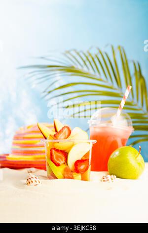 Resort lifestyle - picnic with cold cocktail, fresh tropical fruit salad, sun hat on white sand with palm, sun glare in blue sea Stock Photo