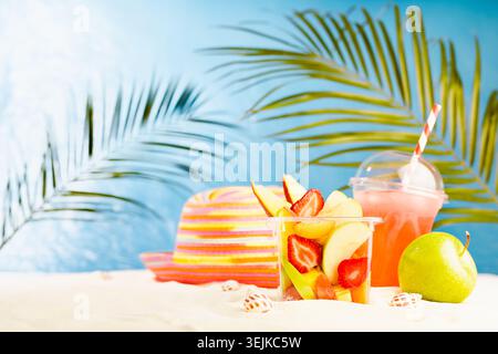 Resort lifestyle - picnic with cold cocktail, fresh tropical fruit salad, sun hat on white sand with palm, sun glare in blue sea Stock Photo