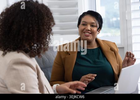 African American female colleagues having one-on-one meeting on couch in lounge using silver laptop. Professional, collaboration, women, jacket, devic Stock Photo