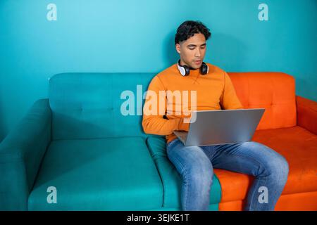 Man sitting on two-tone sofa wearing orange sweatshirt with headphones using laptop, copy space. Teal, couch, home, work, computer, focus, productivit Stock Photo