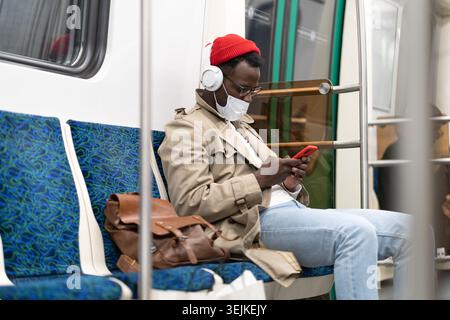 African hipster man in subway train wear face mask using cellphone listens to music with headphones. Stock Photo