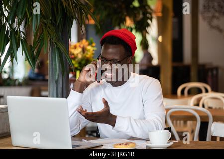 Happy millennial Black man laughing, speaking with friend on mobile phone, remote work in cafe. Stock Photo