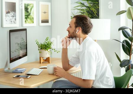 Thoughtful man looking at computer screen working from home remotely. Freelancer guy at workplace with cup of coffee. Male web designer or programmer Stock Photo