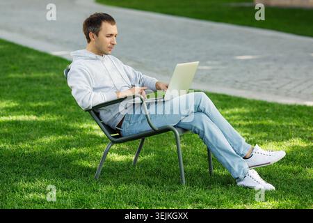 Freelancer man with laptop working in garden, sitting in chair on grass outdoors. Young blogger male work on computer in public park, processes video Stock Photo