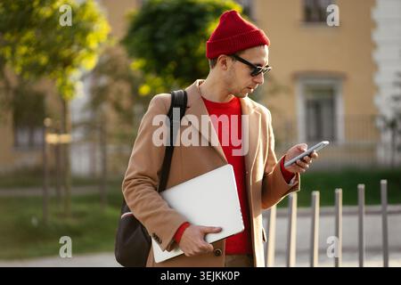 Hipster freelancer, young businessman use smartphone outdoors with laptop in hand walk through park Stock Photo