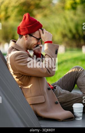 Happy emotional young man has mobile phone call with cheerful smile sit outdoors on bench in park Stock Photo