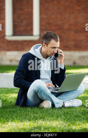Focused freelancer man sitting on grass with legs crossed remote work on laptop, talk on cellphone. Stock Photo