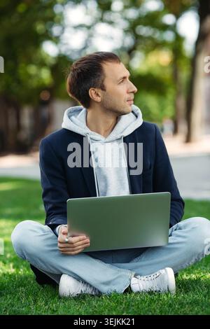 Casual freelance web developer with laptop sit on green grass in park working remotely out of office Stock Photo