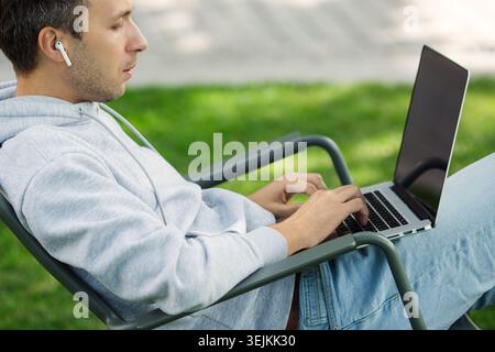 Thoughtful programmer man with laptop working in garden sit in chair on grass outdoors. Freelancer blogger male work on computer in public park proces Stock Photo
