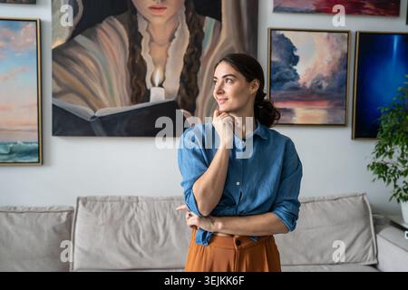 Successful woman painter thinking and getting inspiration while posing against painting exhibition. Stock Photo
