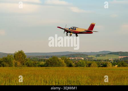 A small red and white monoplane flying low over rural green fields. Stock Photo