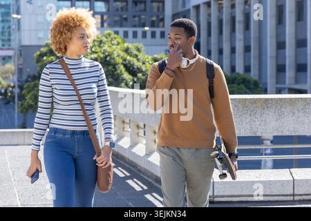African American friends walking on concrete bridge in city, holding phone and carrying skateboard. Duo, urban, sunny, stroll, s Stock Photo