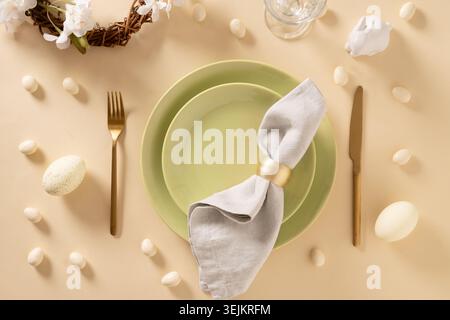 Easter table setting with green plates, bunny, painted eggs on beige background. View from above. Happy Easter celebration. Stock Photo