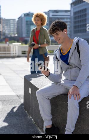 Diverse pair, teen man sitting on plaza bench wearing tracksuit, using smartphone, copy space. Urban, outdoor, sunny, pedestrian, earbuds, backpack, m Stock Photo