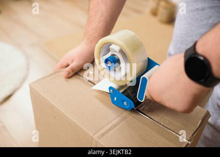 Close-up of man hands using tape gun dispenser to seal cardboard box for moving Stock Photo